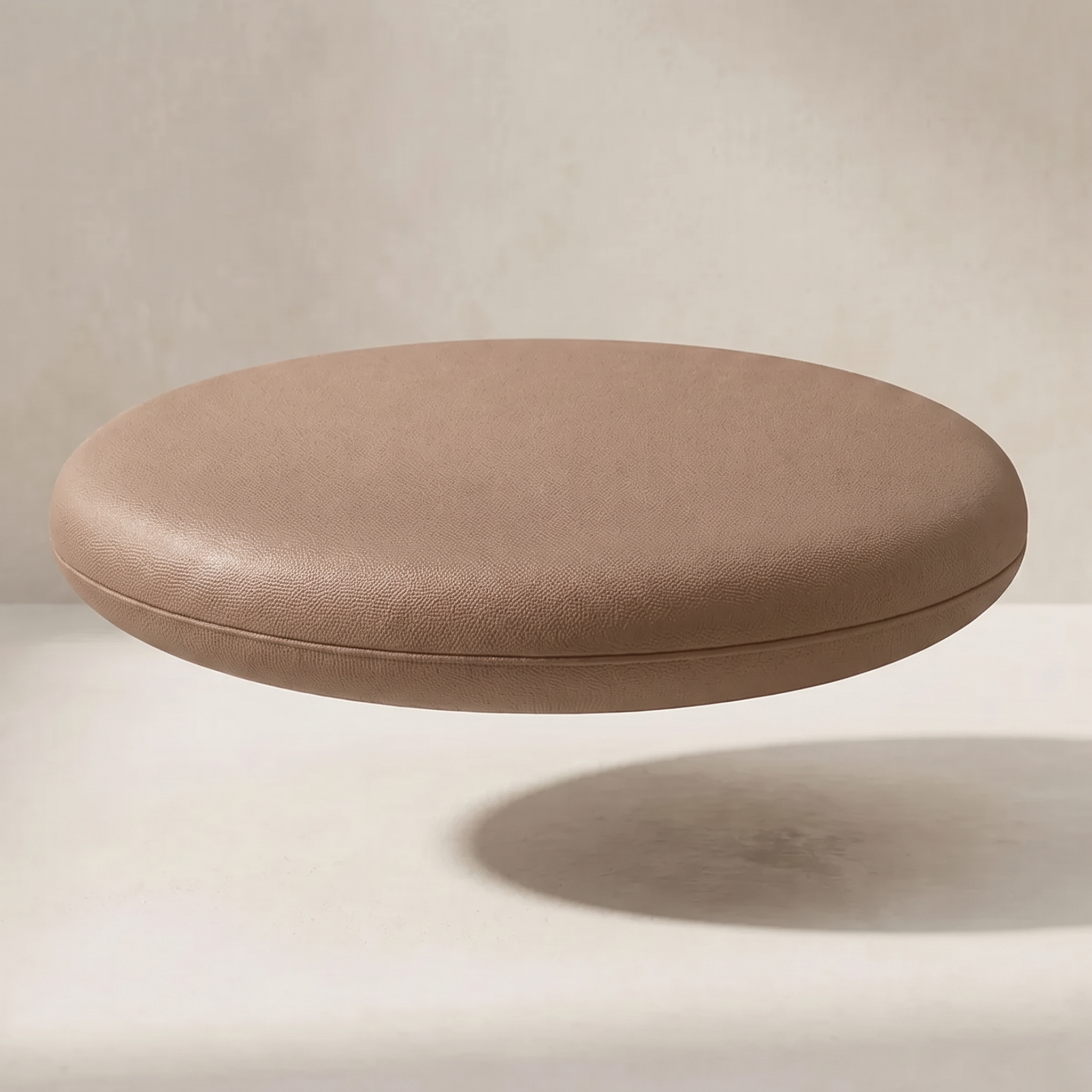A round, low-profile upholstered ottoman or stool with a smooth, pebbled leather-like finish in a warm taupe-brown color, shown floating against a neutral beige background with a soft shadow underneath.