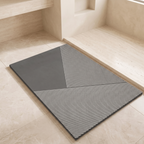 A rectangular gray mat with a geometric pattern of solid dark gray and white diagonal stripes, placed on a light beige stone floor in a minimalist interior setting.
