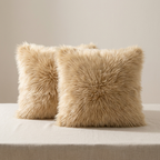 Two fluffy faux-fur throw pillows in a warm beige/tan color, styled on a light beige fabric-covered surface against a neutral beige background.