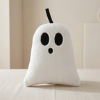 A white plush ghost pillow with black embroidered eyes, nose, and mouth, sitting on a beige fabric surface against a soft beige background.
