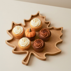 A leaf-shaped wooden serving tray holding six cupcakes, including three white-frosted, two chocolate-frosted, and one small pumpkin-shaped cupcake, on a light beige surface.