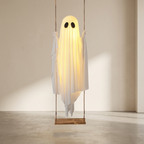 A ghost-shaped hanging lamp or decor piece made from translucent white fabric, glowing warm yellow from within, with black oval eyes and suspended from a wooden swing in a minimalist indoor room.
