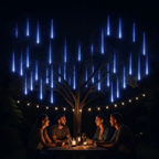 A nighttime outdoor scene with four people seated around a small table under a tree, lit by warm string lights and candles. Above them, many bright blue vertical light streaks resembling falling rain or shooting stars are displayed against a dark sky.