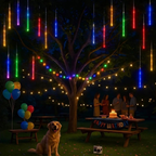 Outdoor birthday party scene at night under a decorated tree with hanging multicolor LED rainbow lights, string lights, balloons, a picnic table with a birthday cake and banner, and a golden retriever sitting in the foreground.