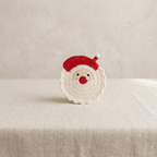 A crocheted Santa Claus face ornament or coaster sits on a textured beige fabric surface against a matching beige background.