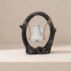 A small decorative figurine of a white ghost sitting on a swing attached to a dark, tree-like circular frame, displayed on a light beige surface against a matching beige background.