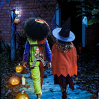 Two children dressed in Halloween costumes walking away on a spooky brick path at night. One child wears a colorful clown-style outfit with a large black afro wig, and the other wears a witch hat and orange cape.