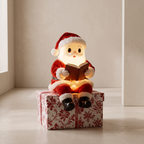 A Santa Claus figurine wearing a red suit and hat, sitting on a red-and-white patterned gift box and reading a glowing book. The scene is photographed indoors against a warm beige wall and floor.