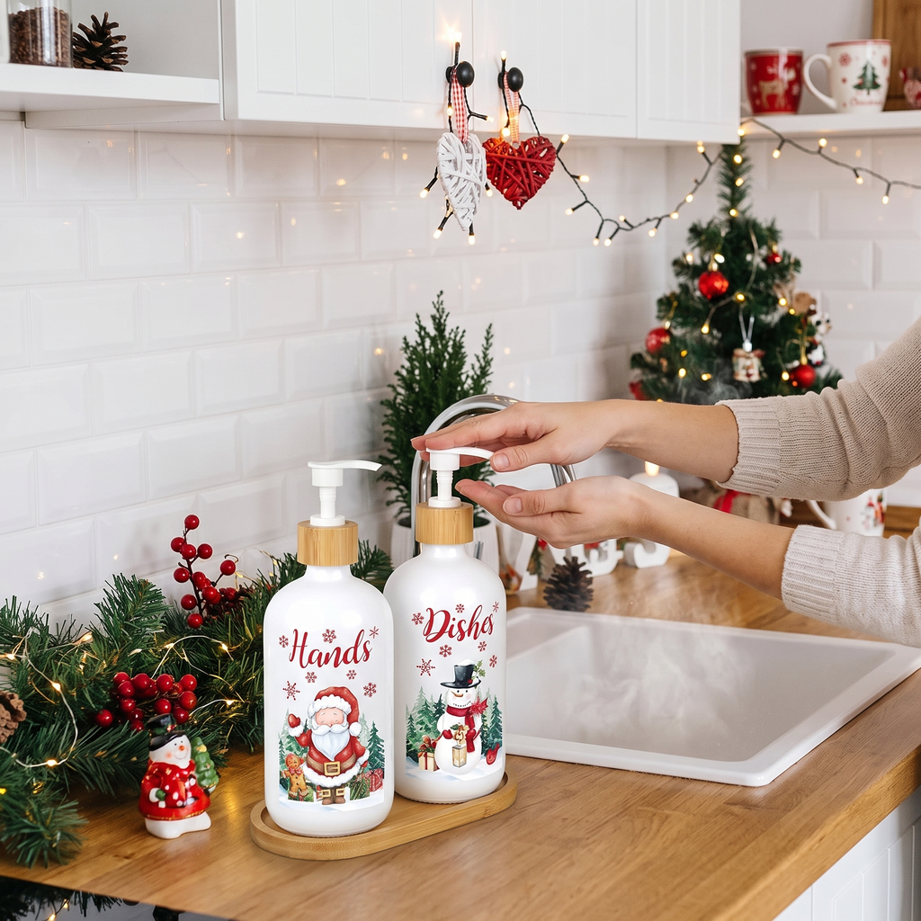 Two white Christmas-themed pump bottles for dishwater and hand sanitizer with wooden caps, shown on a kitchen counter with a person's hands using one. The background features a decorated Christmas tree, shelves, and warm string lights.