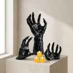 Three black sculptural zombie hands arranged on a light stone pedestal, with three small yellow tea light candles in front, set against a warm beige wall near a window.