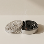 A round metal incense holder with a matching lid, shown open with a black coil of incense inside and a small red ember. The product is photographed on a clean, light beige surface against a matching beige wall.