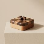 A small rectangular wooden keepsake box with a checkered dark and light wood lid and a matching bow on top, displayed on a beige pedestal against a warm beige background.