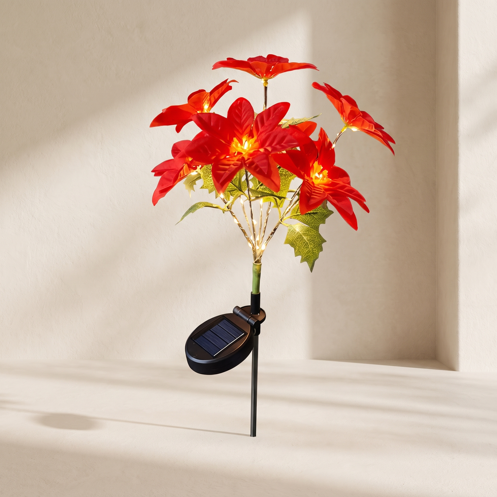 A solar-powered artificial poinsettia plant with glowing red flowers and green leaves, mounted on a black stake with an attached oval solar panel, set against a warm beige wall with soft shadows.