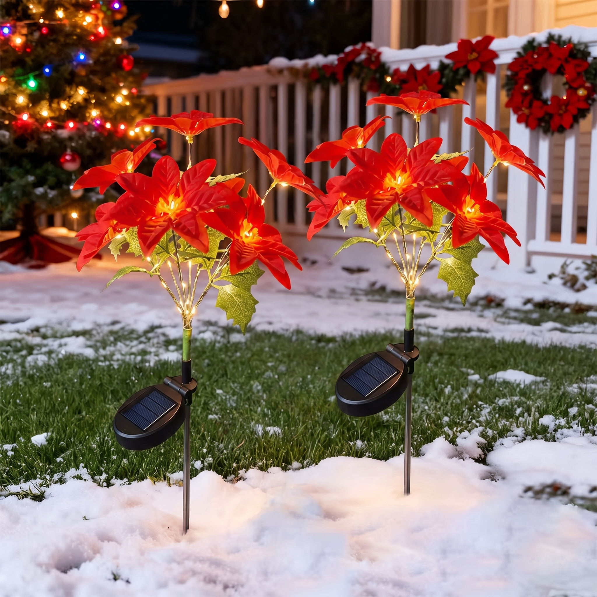 Two solar-powered artificial poinsettia garden lights with red flowers, green leaves, and warm white string lights, planted in snow on a lawn in front of a white fence and decorated Christmas tree.