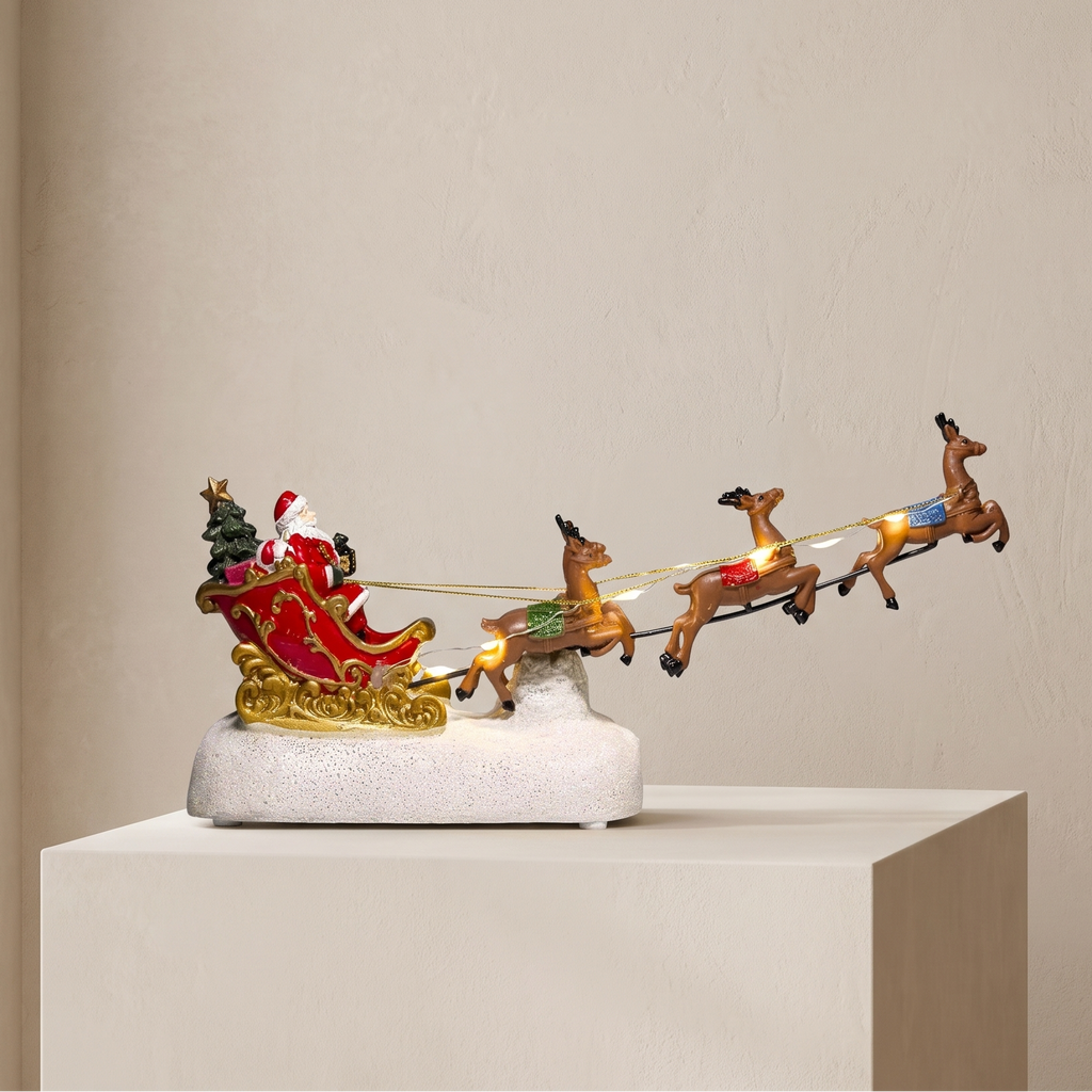 A Santa Claus sleigh decoration displayed on a white pedestal in a minimalist room. The ornament features Santa in a red sleigh with gold trim, pulled by four brown reindeer with glowing amber eyes, all mounted on a white textured base.