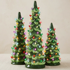 Three green ceramic Christmas tree ornaments with colorful lit bulbs, displayed on a neutral beige surface against a matching background.