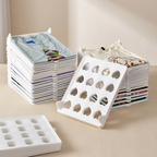 White plastic t-shirt stacking and folding trays shown with several stacks of folded shirts in the background on a beige tabletop.