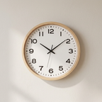 A minimalist analog wall clock with a light natural wood frame, a white face, and black numerals and hands, mounted on a neutral off-white wall.