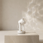 A modern white adjustable spotlight lamp with a clear glass shade, resting on a light beige stone pedestal. The lamp is turned on, casting a soft, rippled water-like shadow pattern on the beige wall behind it.