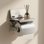 A wall-mounted stainless steel toilet paper holder with a small shelf holding a diffuser and a smartphone, shown against a beige wall with sunlight and shadows.