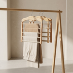 A wooden trouser rack with three hangers holding folded beige and off-white trousers, set against a minimalist beige wall and floor.