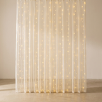 A floor-length sheer curtain panel draped in soft vertical folds, illuminated with warm white string lights that glow against a neutral beige wall and floor.
