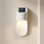 A modern white solar wall light mounted on a beige wall, featuring a small dark solar panel on a circular top cap above a glowing warm white light housing.