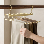 A gold metal multi-hanger rod hanging from a dark wooden rail, holding several pairs of folded pants in neutral and brown tones. A person's hand is visible adjusting the pants.