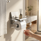 A wall-mounted stainless steel toilet paper holder with a roll of toilet paper, shown in a bright modern bathroom. A hand is pulling the toilet paper, and a white towel hangs on the left.