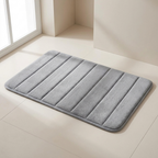 A rectangular gray bath mat with vertical channel tufting, shown on a light beige tiled floor in a minimalist bathroom setting.