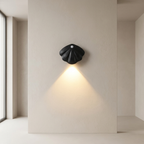 A black sculptural wall sconce with a shell-like form, mounted on a beige plaster wall and casting a warm downward light in a minimalist interior setting.