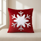 A square red throw pillow with a large white snowflake design on the front, resting on a light beige upholstered bench in a softly lit room.