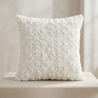 A square white textured throw pillow with a raised floral or snowflake-like pattern, resting on a beige upholstered surface against a soft neutral background.