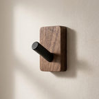 A wall-mounted coat rack hook with a rectangular dark wood backplate and a cylindrical black metal hook, shown against a warm off-white textured wall.