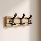 A three-hook wooden coat rack with black metal hooks mounted on a light natural wood plank, shown against a plain off-white wall.