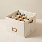 An off-white fabric storage box with a grid of compartments holding ribbon spools, featuring brown handles and a label slot on the front, set against a neutral beige background.