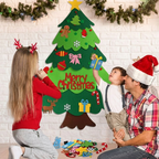 A family of three decorating a large felt Christmas tree wall hanging with colorful ornaments, gifts, and candy canes. The girl wears red reindeer antlers, the boy wears a Santa hat, and the man wears a Santa hat and plaid shirt.