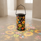 A black cylindrical lantern with a wire handle and a colorful floral print, placed on a concrete floor that projects a sunflower and butterfly pattern. The lantern is shown in a minimalist, modern interior setting.