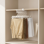A white multi-hanger closet rod hanging in a beige wardrobe, holding several pairs of folded pants and shorts in neutral tones including tan, brown, and white.