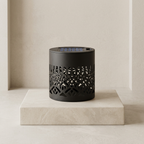 A black cylindrical solar lantern with an intricate laser-cut geometric pattern on the body, displayed on a light beige stone pedestal against a matching beige textured wall.