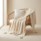 A cream-colored textured knit throw blanket with tassels draped over a woven rattan armchair, paired with a matching cream pillow, set in a warm neutral room.