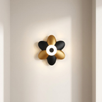 A modern flower-shaped wall sconce light fixture with alternating matte black and brushed gold petals, featuring a glowing white circular center, mounted on a warm beige wall.