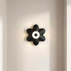 A black flower-shaped wall sconce with a glowing white circular center, mounted on a warm off-white wall.