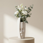 A modern sculptural ceramic vase with a face design, filled with white flowers and green eucalyptus leaves, displayed on a matching round pedestal against a warm beige background.