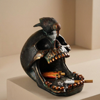 A gothic skull-shaped ashtray with a small crow perched on top, featuring an open mouth filled with cigarette butts and a single lit cigarette, set against a warm beige background.