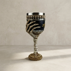 A decorative goblet with a skeletal hand holding the bowl, featuring a dark blue and gold patterned body, a silver rim, and a bone-styled stem and base, photographed against a neutral textured beige background.