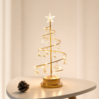 A spiral gold metal Christmas tree centerpiece with clear hanging crystal ornaments and a lit star topper, displayed on a small white table with a pinecone beside it.