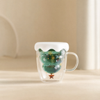 A clear double-walled glass mug with a white lid and a green Christmas tree-shaped tea infuser inside, set on a light beige surface against a soft beige background.