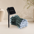 Solar garden string lights with a black solar panel stake and a bundle of green wire lights with white LED bulbs, displayed on a light beige surface.