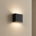 A modern square black wall sconce light fixture mounted on a beige textured wall, casting a warm glow downward and upward.